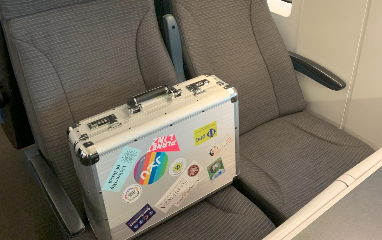 Ein silberner Koffer, der auf einem Sitz in einem Zug steht, und mit vielen Stickern beklebt ist